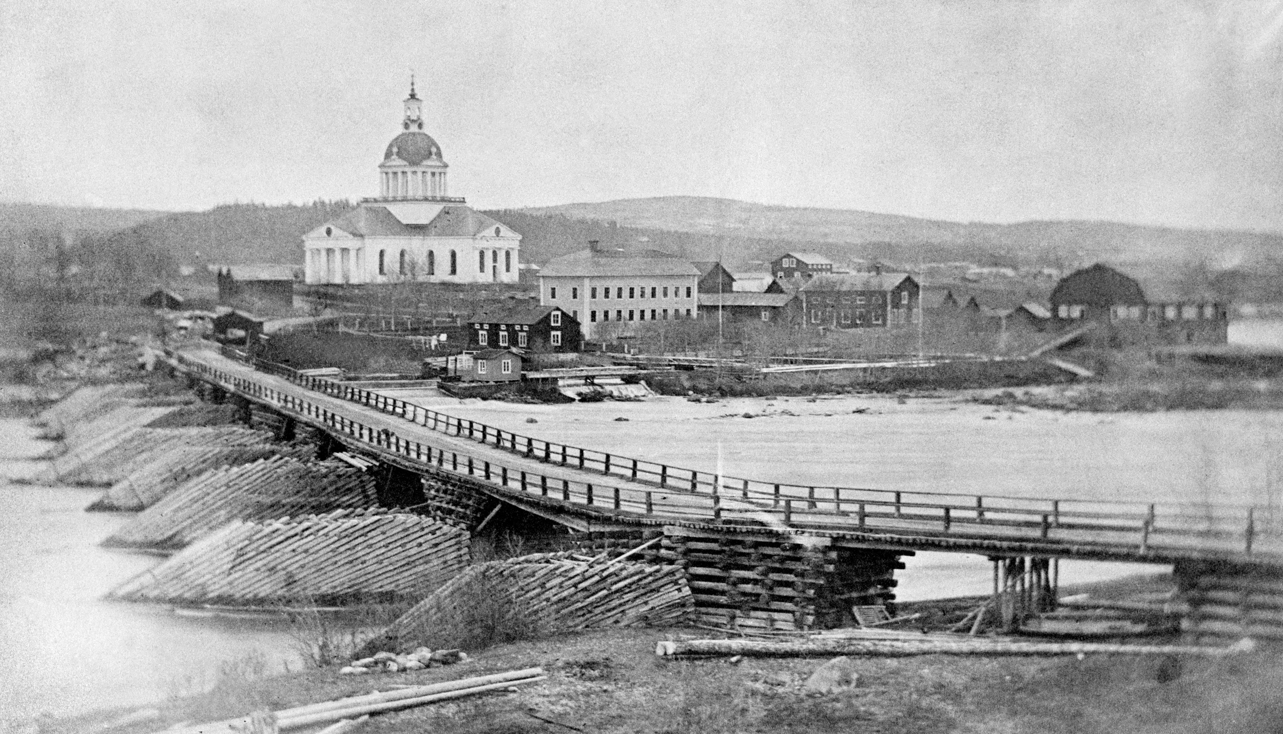 Storbron före 1864. En balkbro med stenfyllda, timrade brokar och stenfyllda isbrytare av timmer. I bakgrunden landskyrkan samt Lejonströms sågverk och herrgård. Foto: Okänd fotograf/Skellefteå museum.