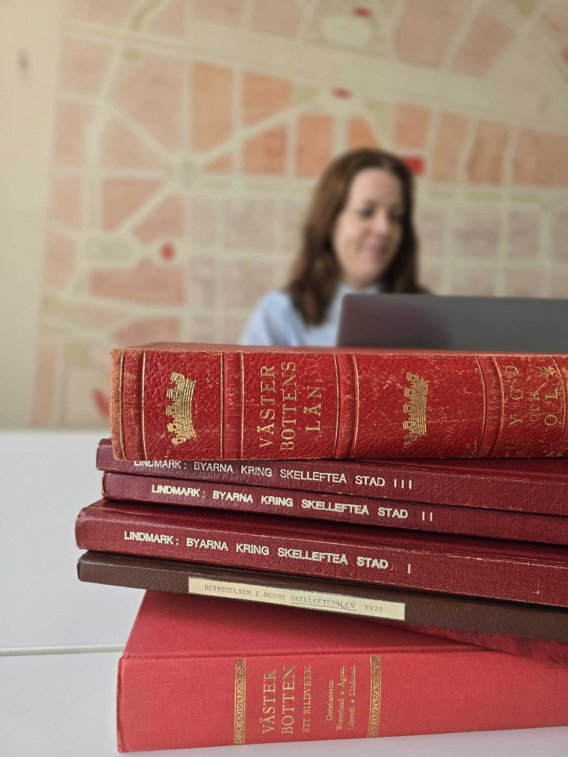 En hög med röda böcker om Västerbotten och Skellefteå i förgrunden. I bakgrunden syns en suddig person som arbetar vid en dator framför en karta.