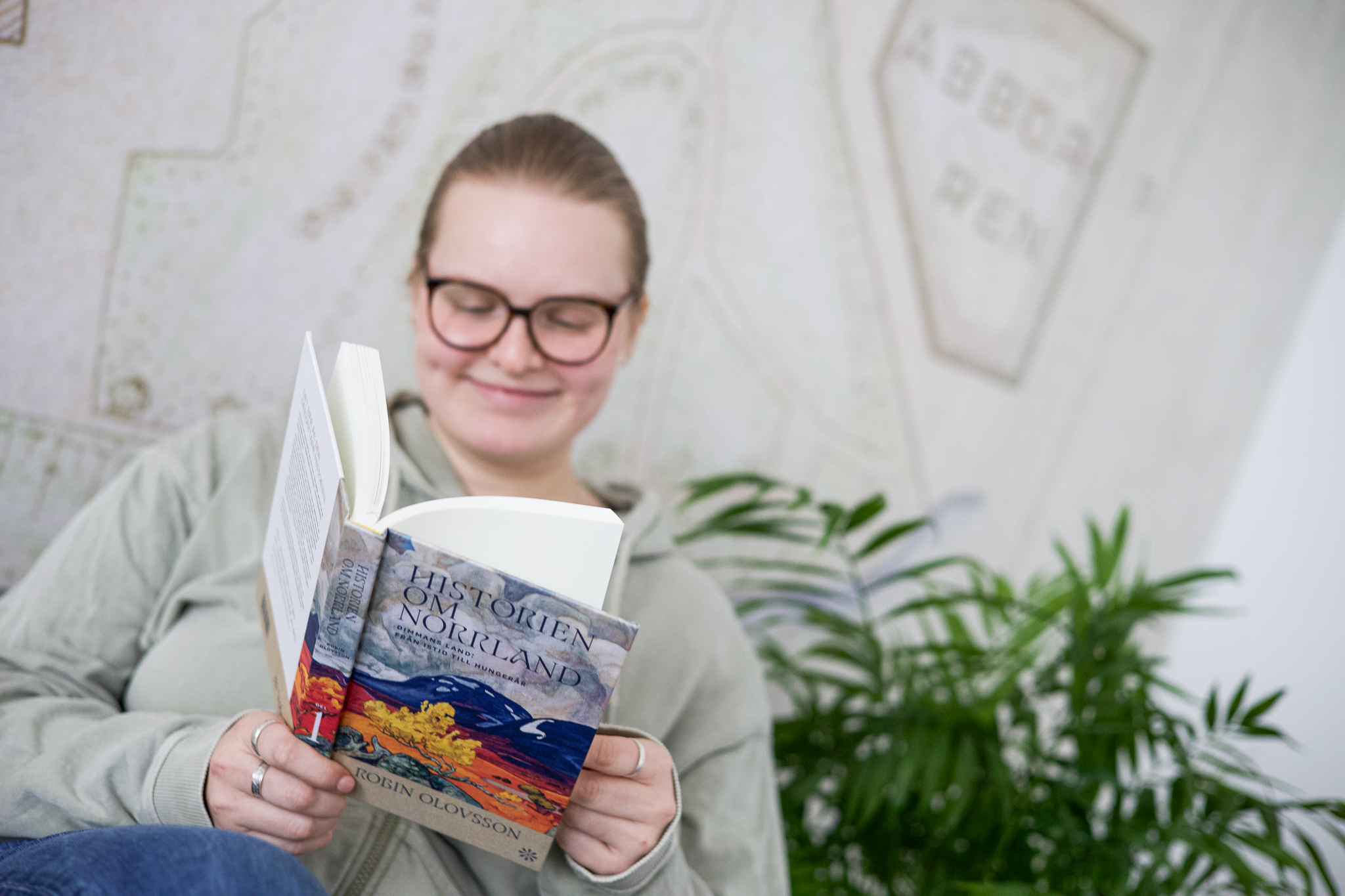 En person sitter och läser boken Historien om Norrland i Läroverket. I bakgrunden syns en vägg med kartmotiv och en grön växt.
